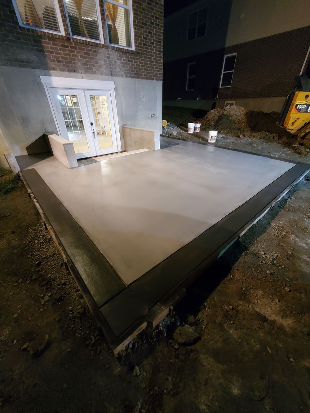 Freshly poured concrete patio beside a brick building at night, with a lit glass door and raised edge formwork.