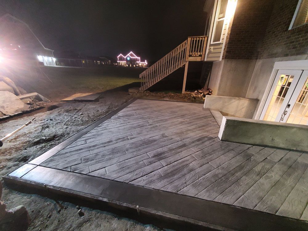 Nighttime wooden boardwalk beside a lit building and dirt slope, with stairs leading up to a deck.