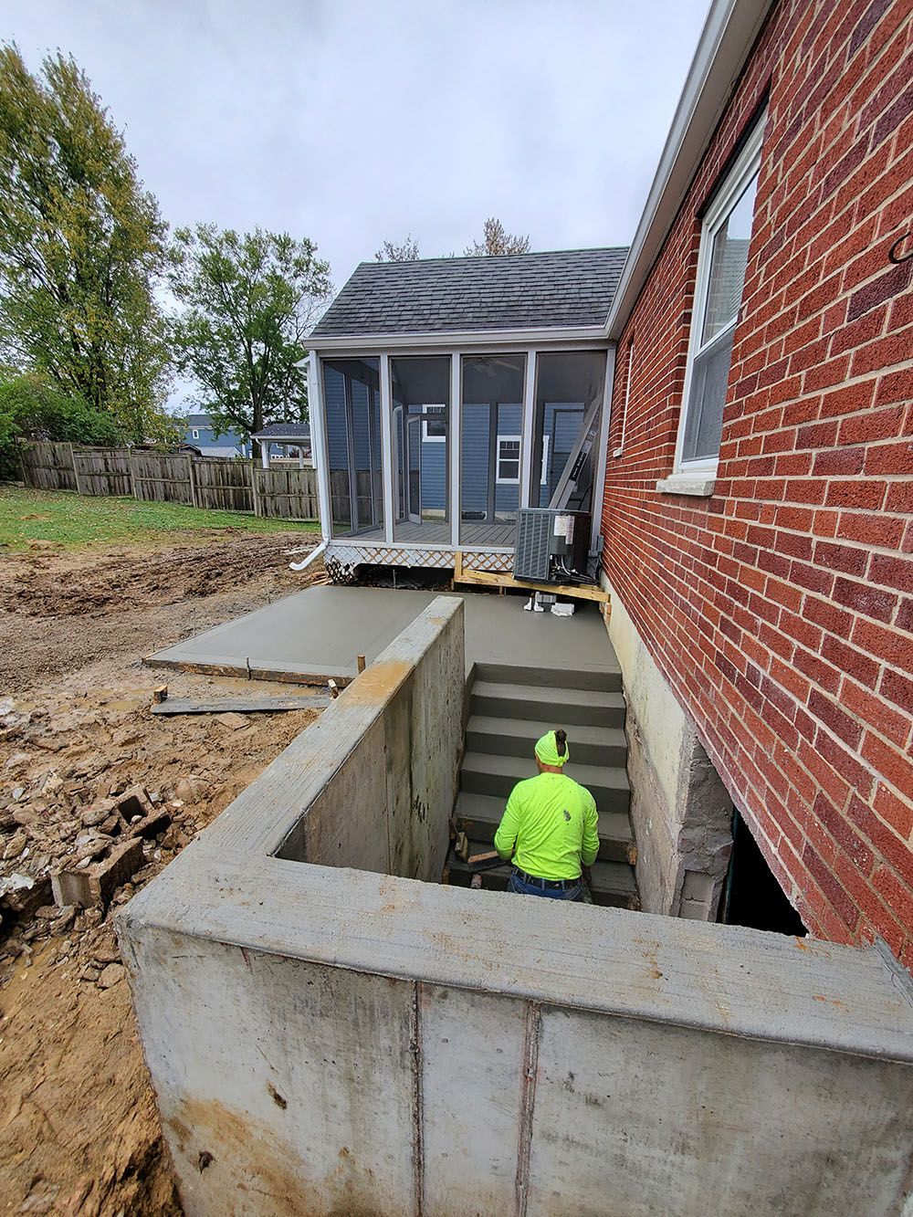 Person in neon jacket in basement excavation beside a brick house and concrete foundation walls