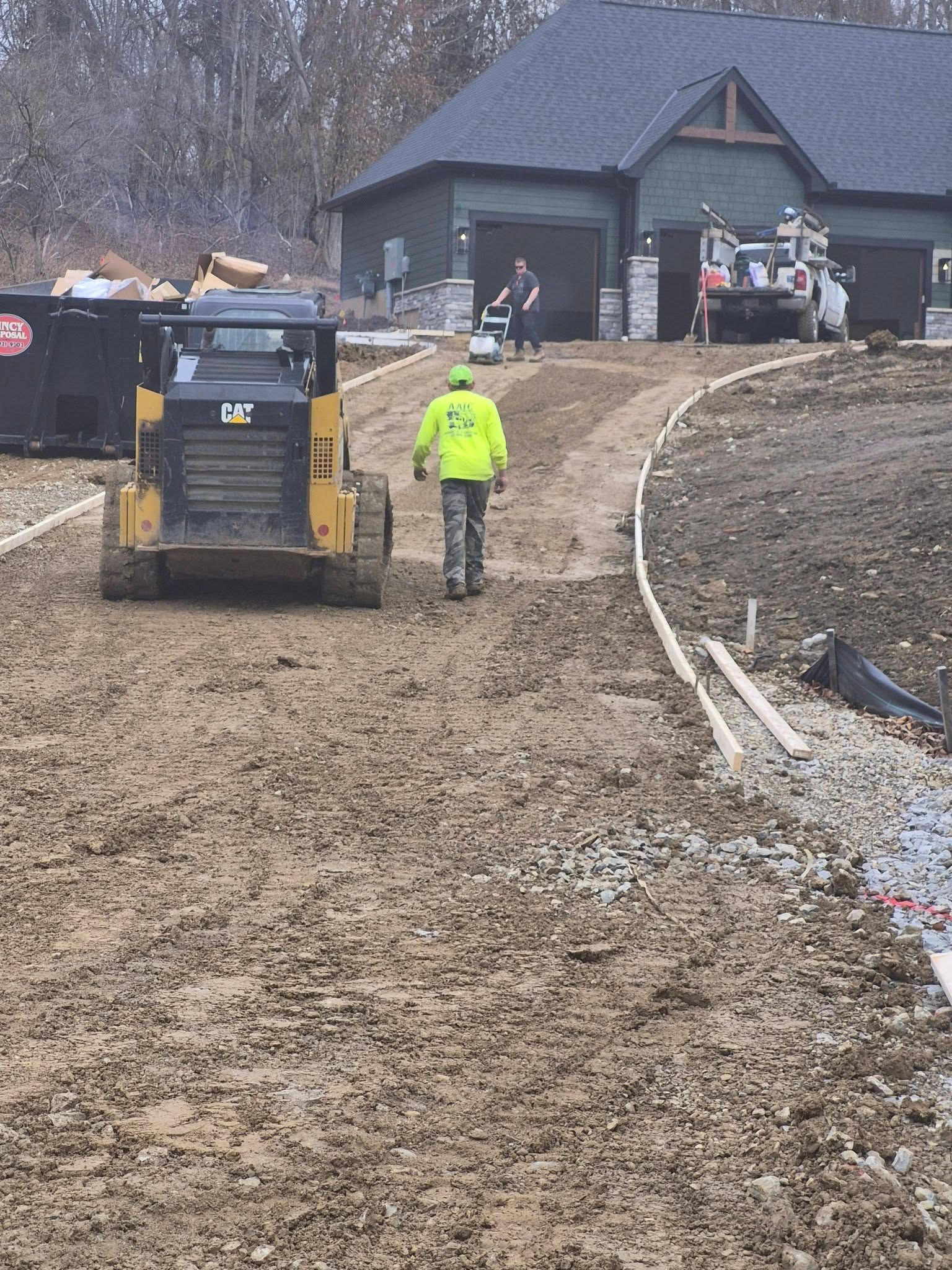Construction worker guiding a skid steer on a muddy driveway near a house under construction