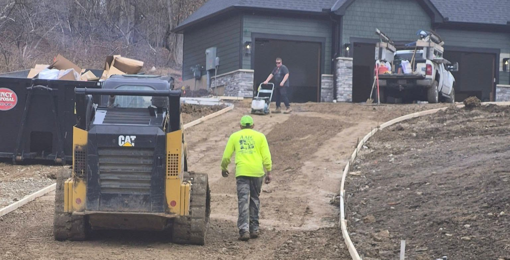 Construction worker guiding a skid steer on a muddy driveway near a house under construction