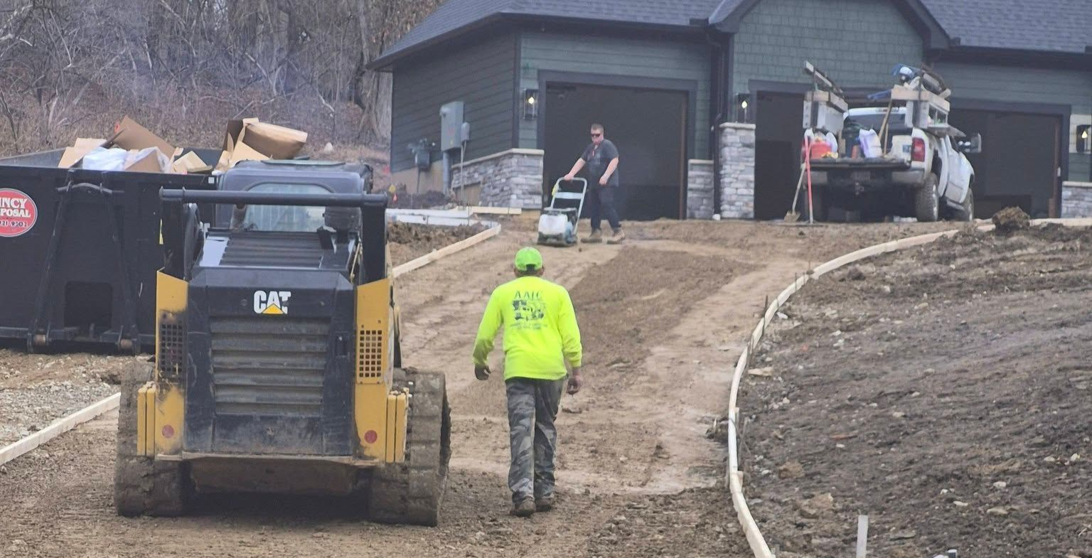 Construction worker guiding a skid steer on a muddy driveway near a house under construction