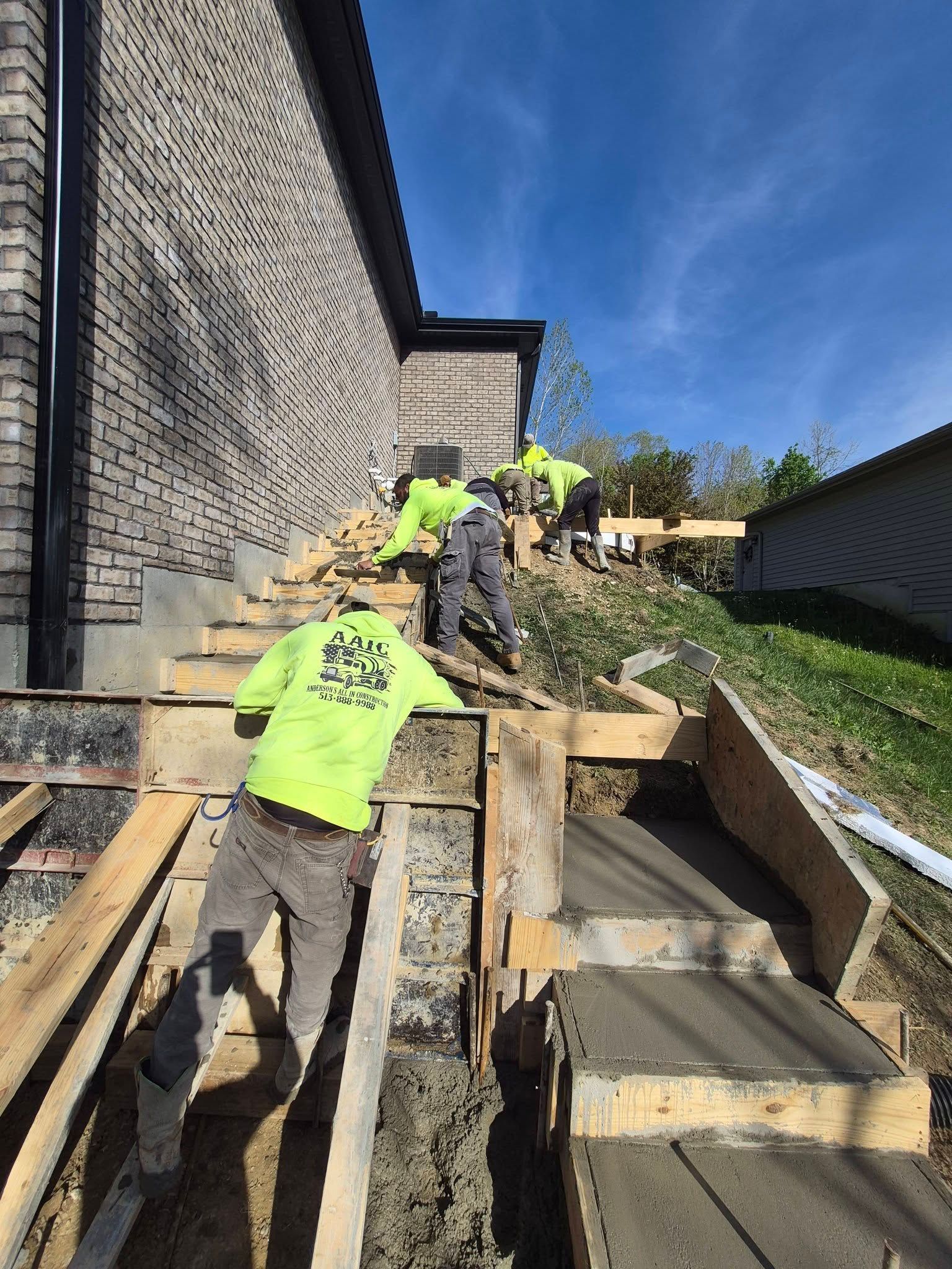 Workers in neon shirts building outdoor wooden stairs beside a stone wall on a sunny day