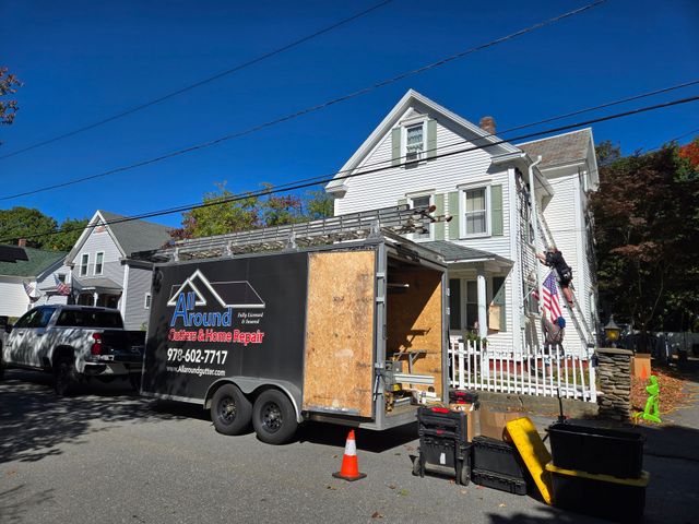 Black utility trailer and parked van outside a white house, with orange cones and an American flag.