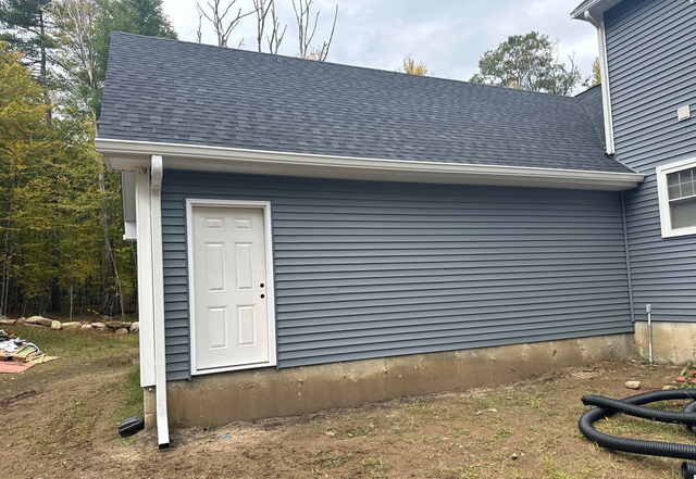 Blue house side with white door, shingled roof, and dirt yard.