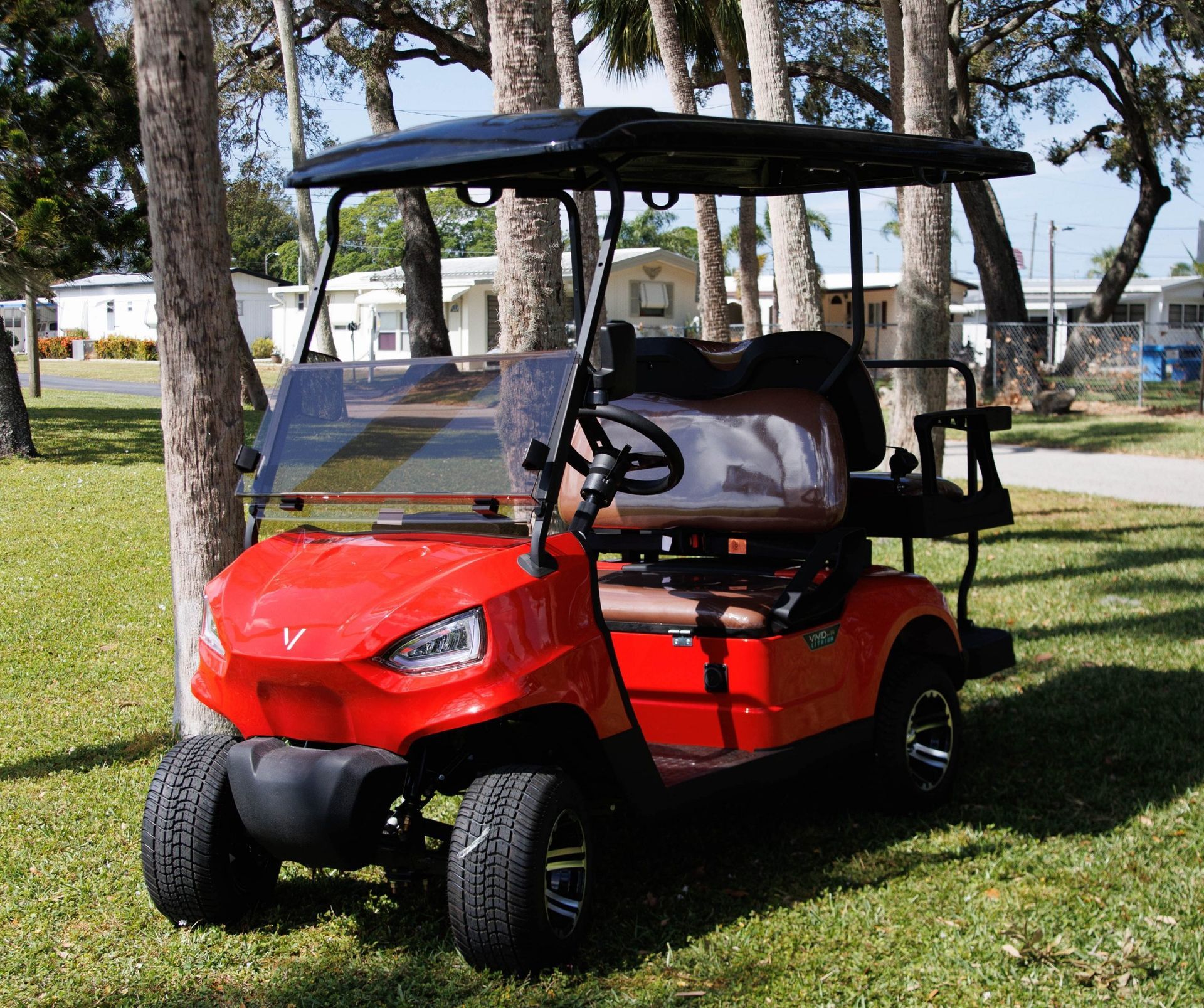 Red golf cart with black roof parked on grass near trees and houses.