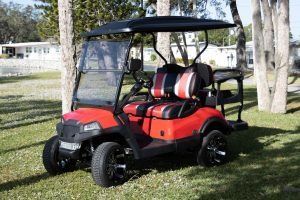 Red and black golf cart on grass with black wheels and canopy; houses in background.