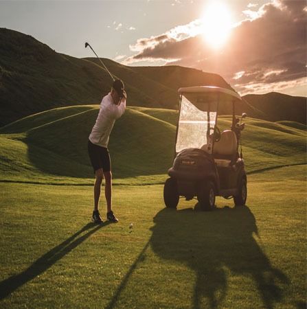 Golfer swings club on green course with cart, sun overhead.