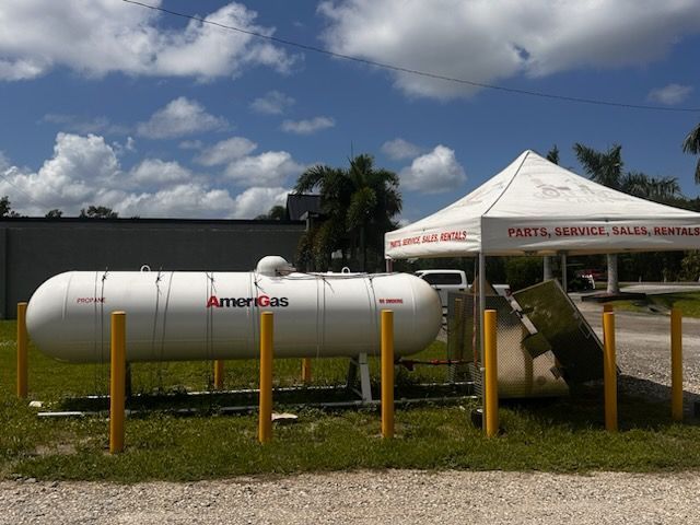 White propane tank beside fenced lot and canopy tent under blue sky