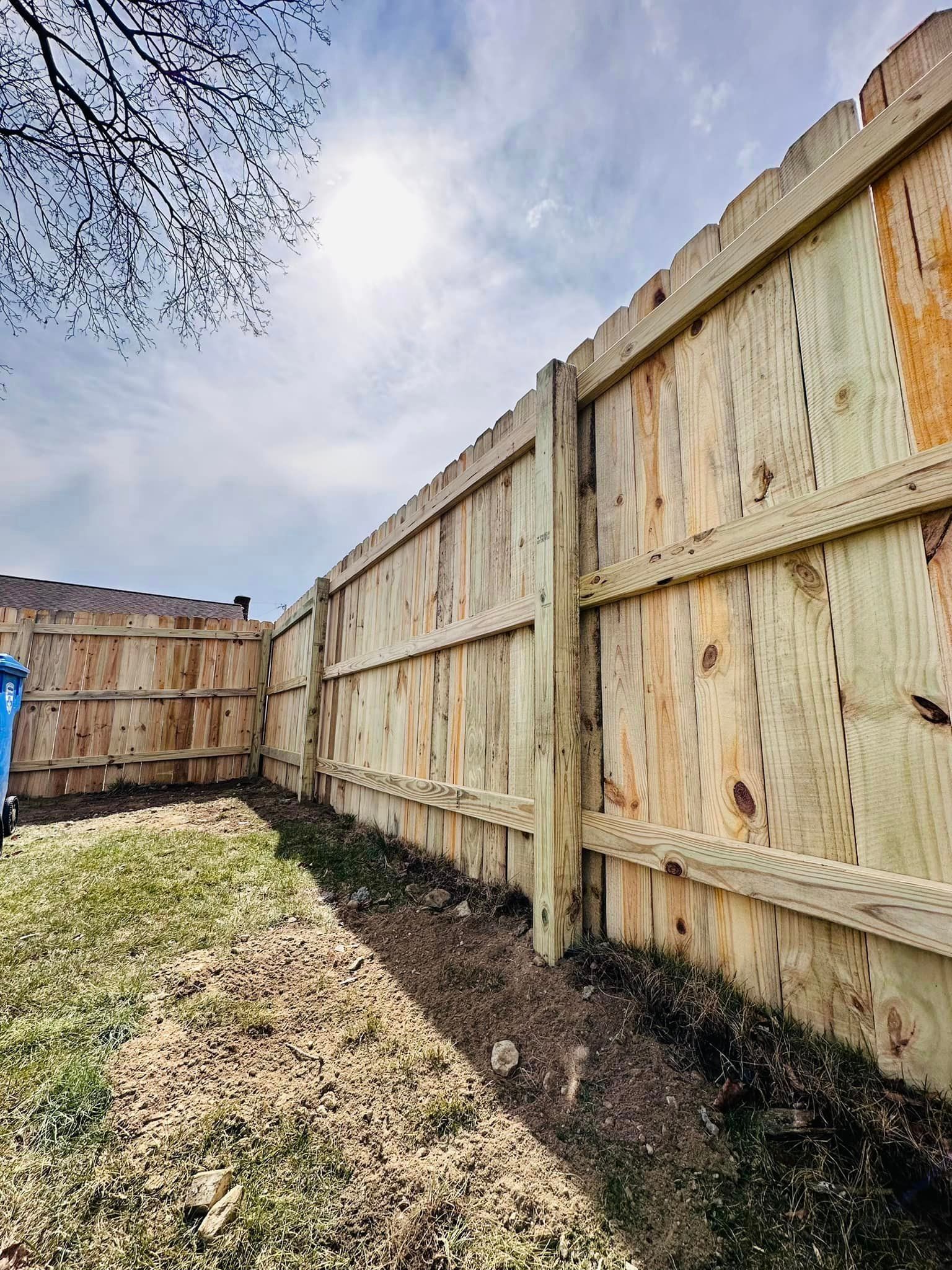 a wooden fence in a backyard with a blue trash can in the background .