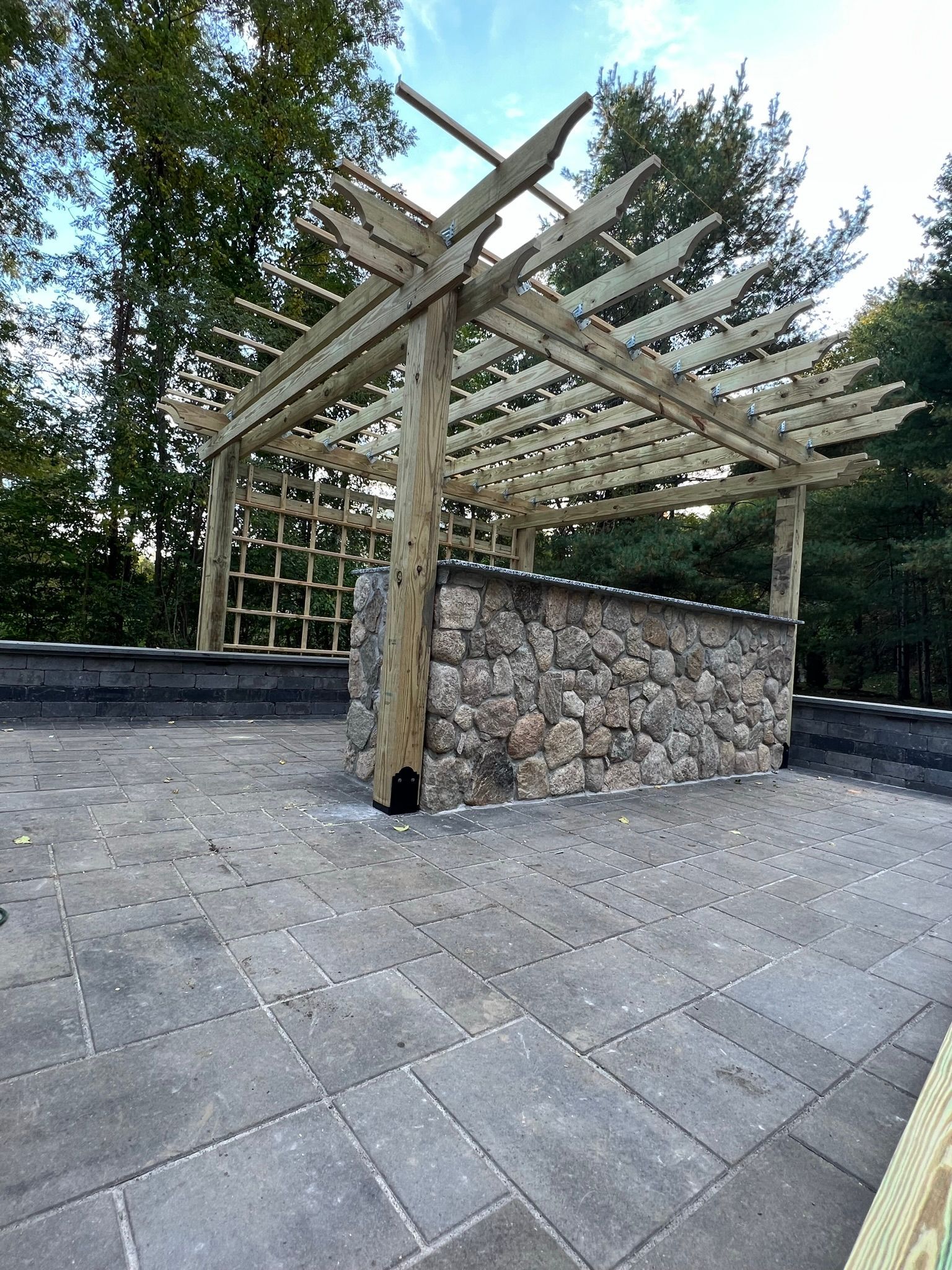 A wooden pergola is sitting on top of a patio next to a stone wall.