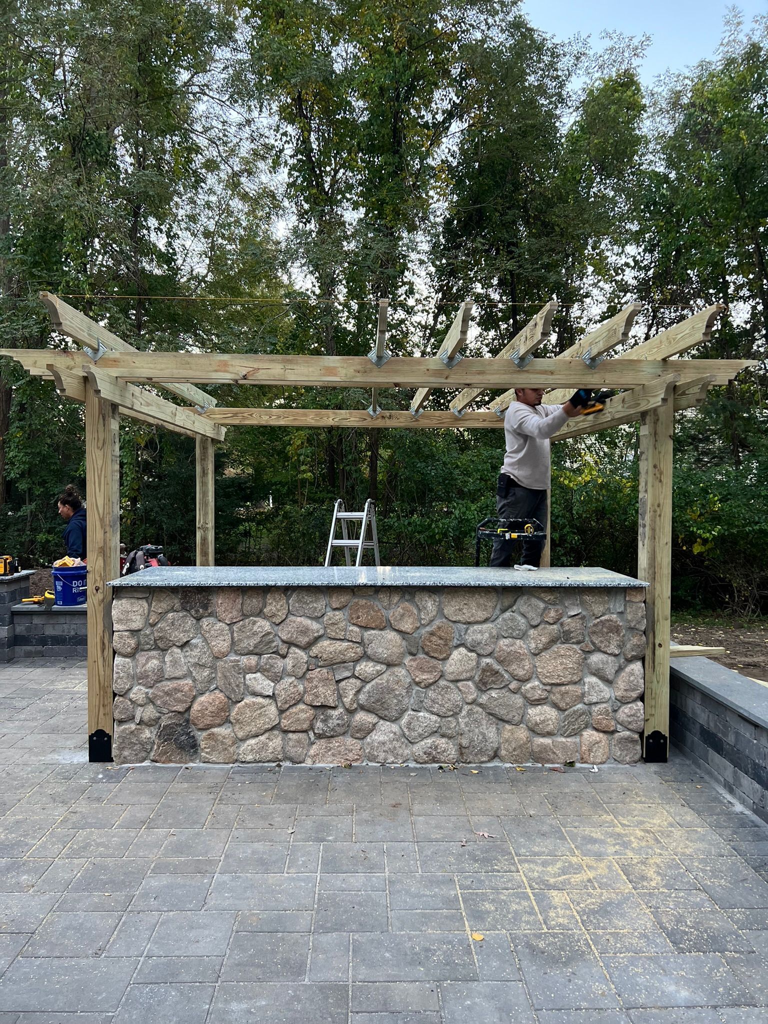 A man is working on a wooden pergola over a stone wall.