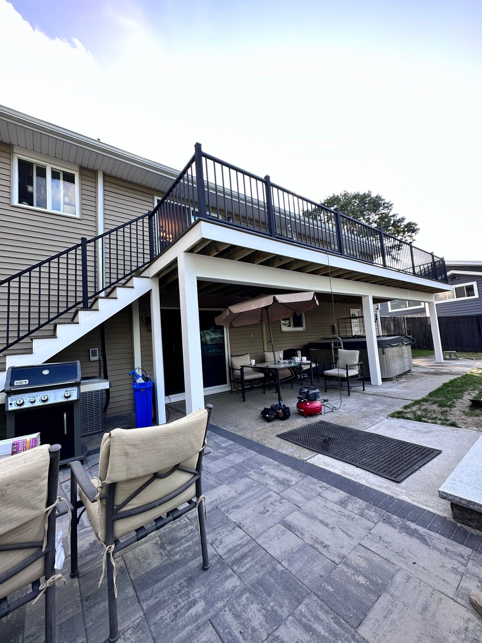 A patio with chairs , umbrellas , a grill and stairs leading up to a deck.