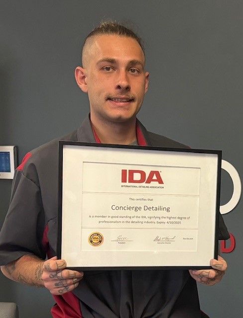 Man holding a framed certificate from IDA, Concierge Detailing. He's smiling, wearing work attire. Gray wall in background.