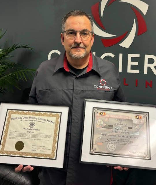 Man in a black uniform holds two framed certificates, smiling, in front of a company logo.