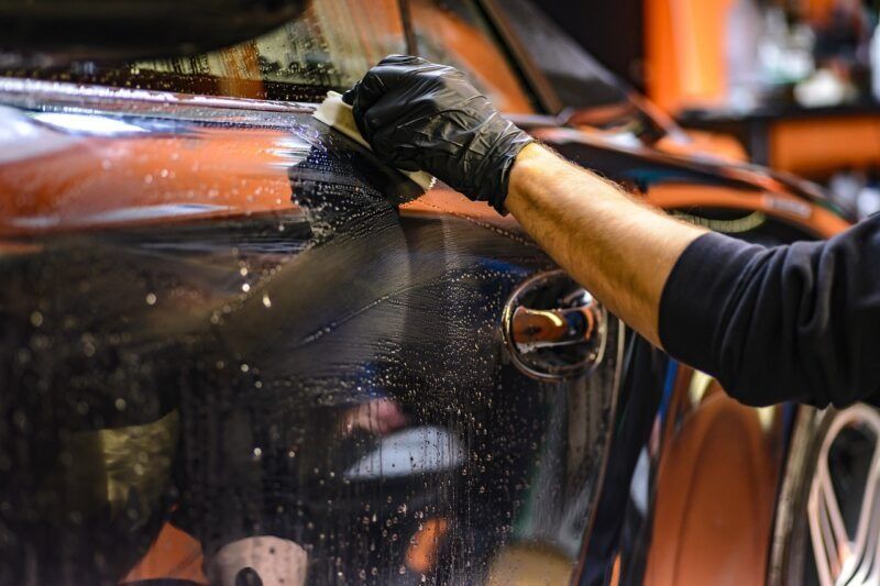 Person washing a black car, wearing black gloves, with a sponge and soapy water.