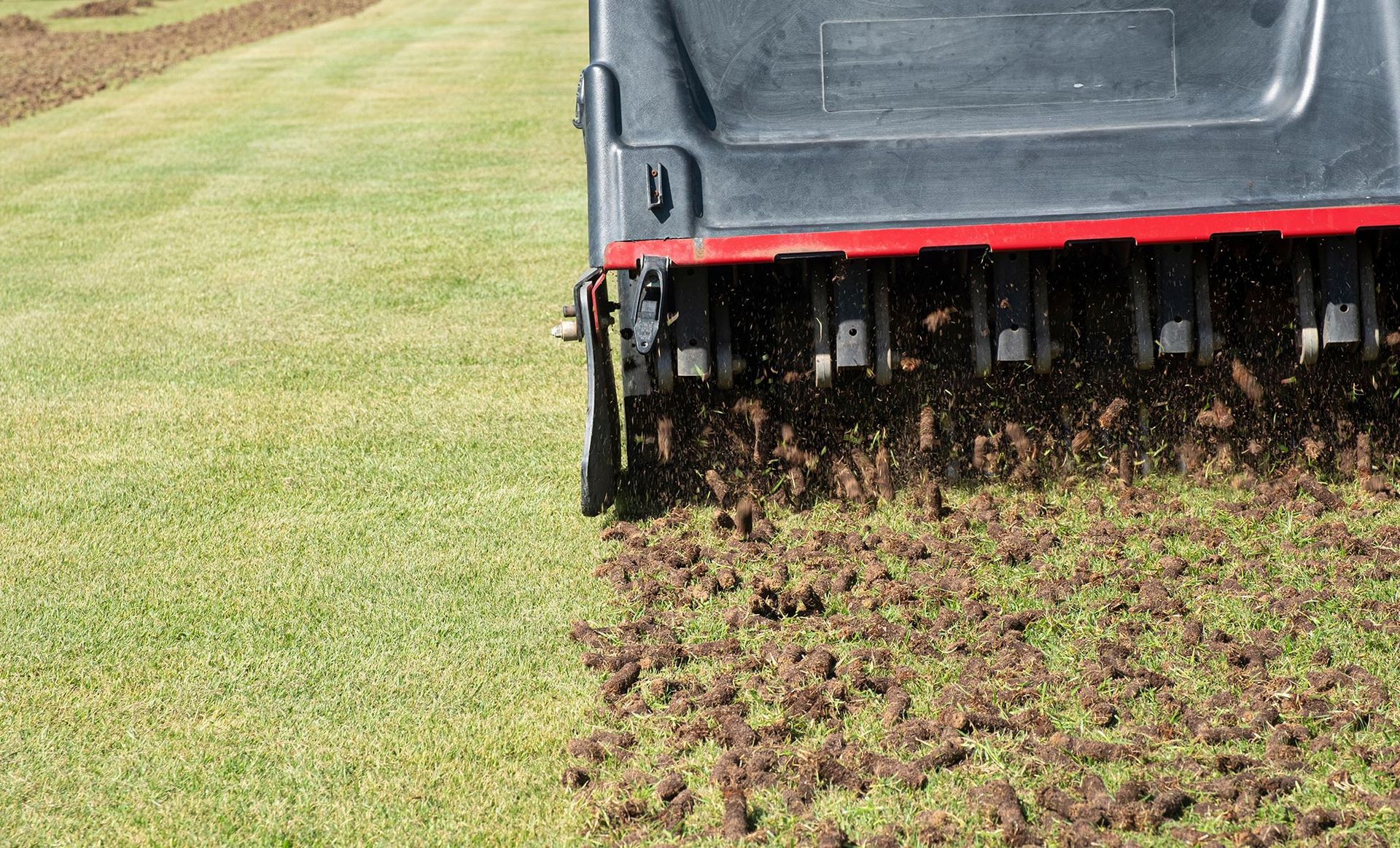 Aerator on a lawn, pulling plugs of soil. The machine is black and red; grass is green.