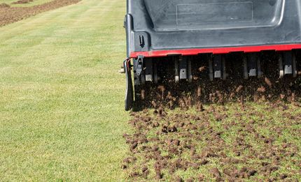 Aerator on a lawn, pulling plugs of soil. The machine is black and red; grass is green.