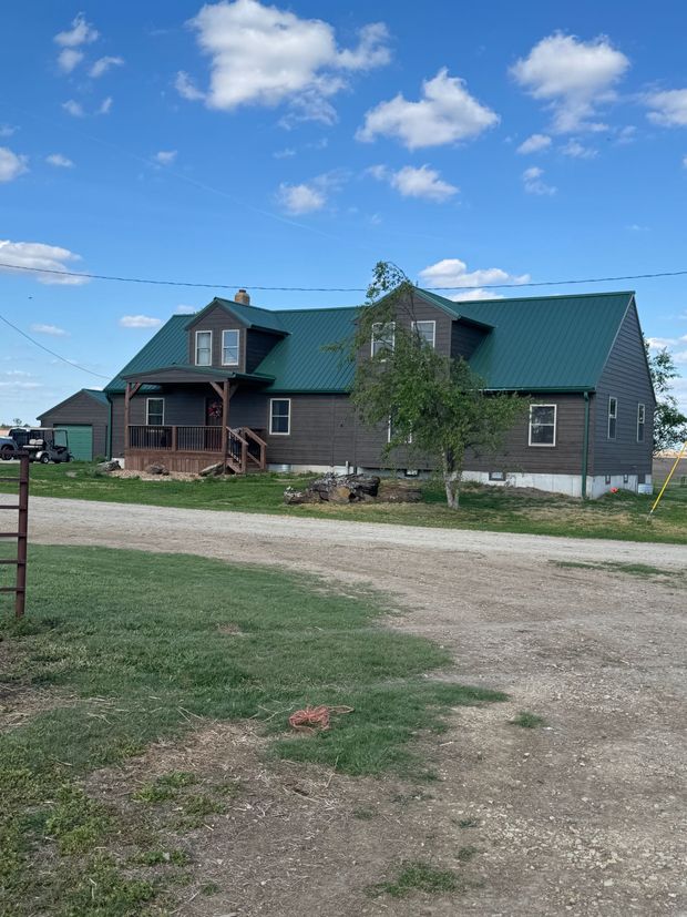 Farmhouse with green roof and gray siding on a gravel driveway under a blue sky.