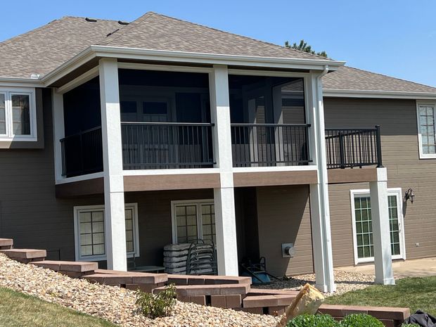 Back of a house with a screened-in porch and a balcony, tan siding, white trim, and a brick retaining wall.
