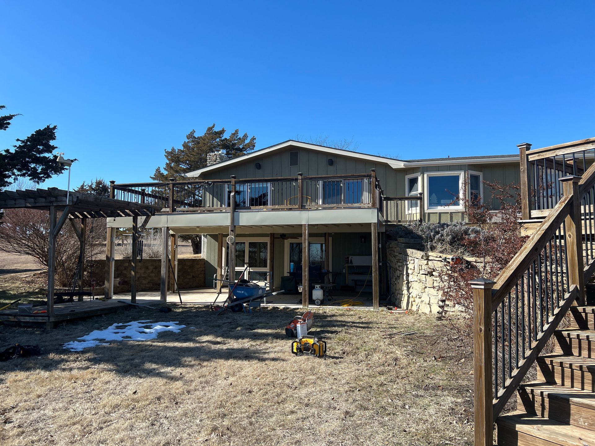 House with a wooden deck, stairs, and pergola. Winter scene with patches of snow on the ground.