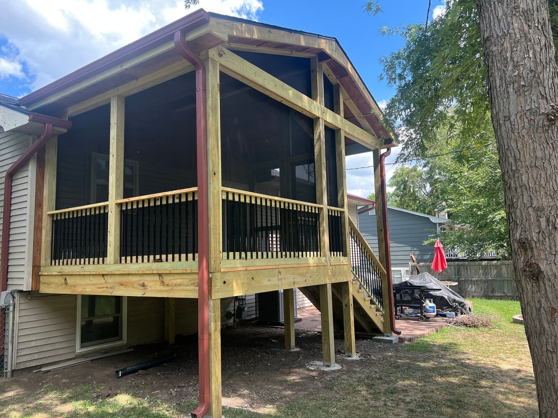 Screened-in porch addition with black railings and steps. Set in a backyard with a tree and partial view of a neighboring house.
