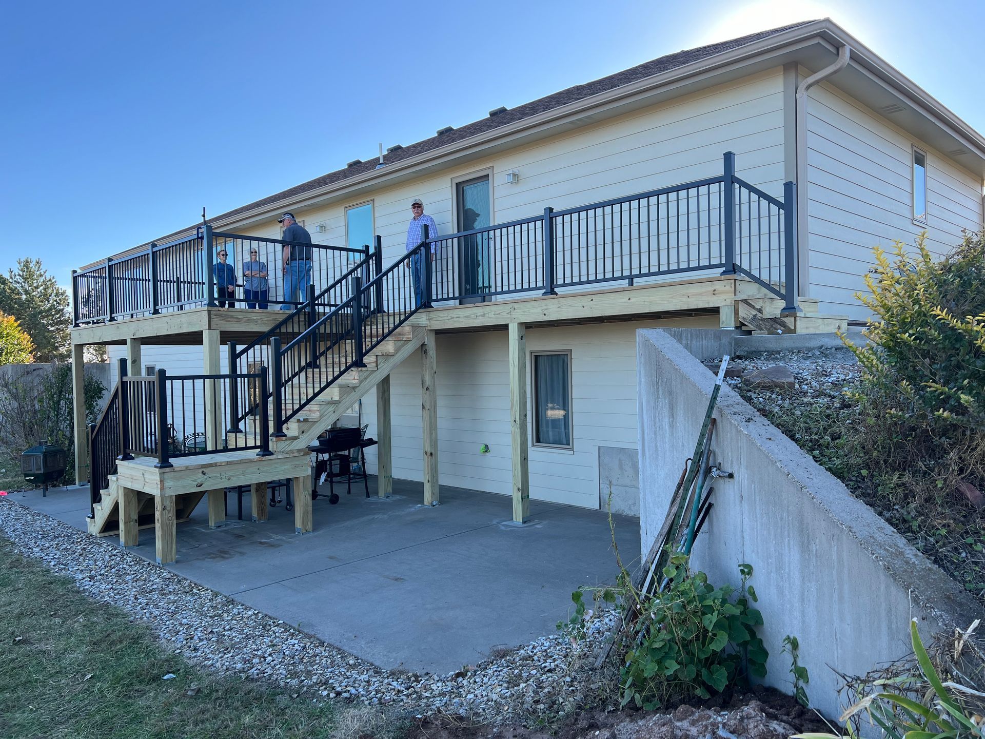Two-story deck with black railings and stairs attached to a beige house, with people standing on the deck.
