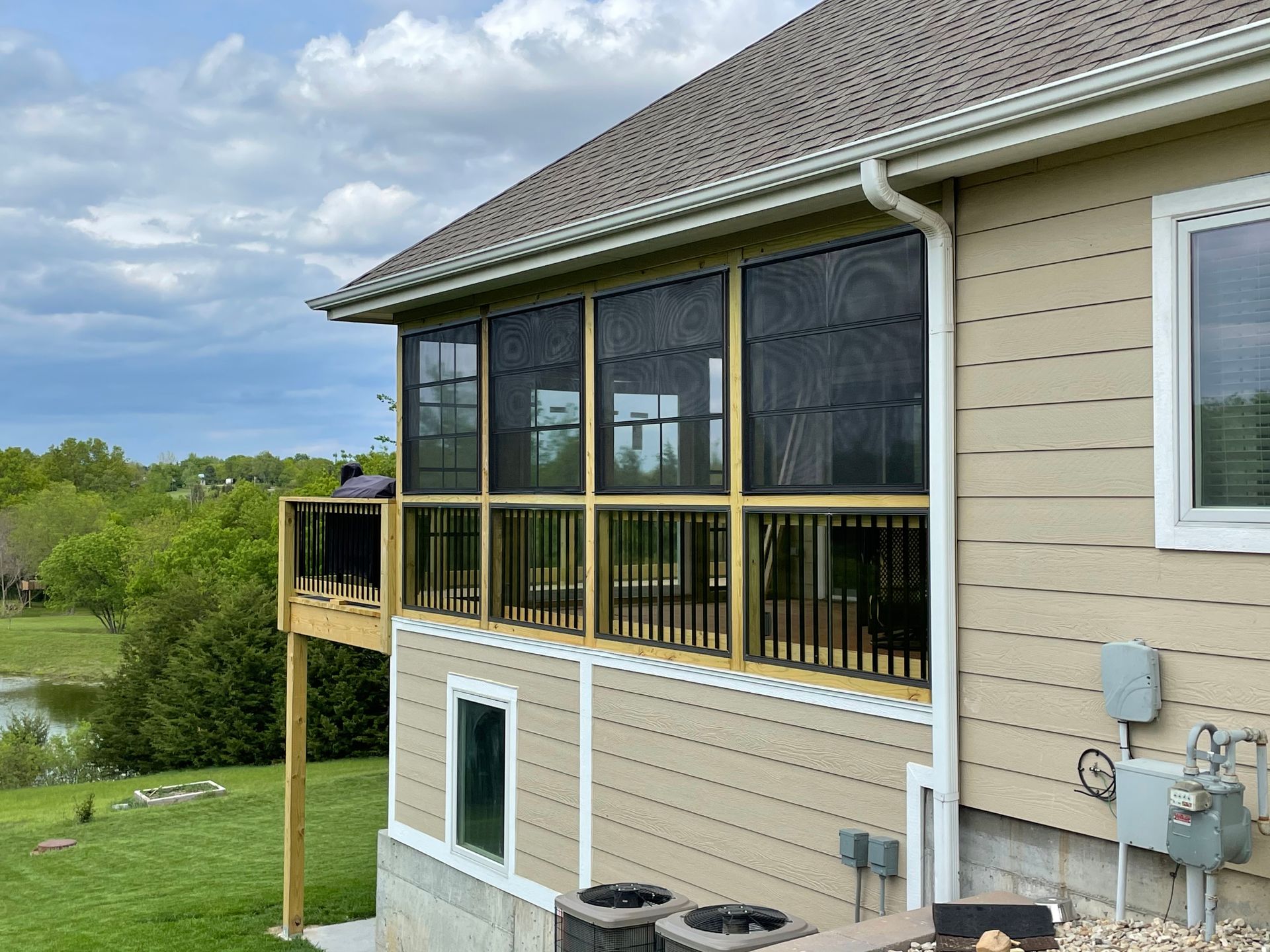 Screened porch with dark screens, beige siding, and a wooden deck overlooking a green landscape and lake.