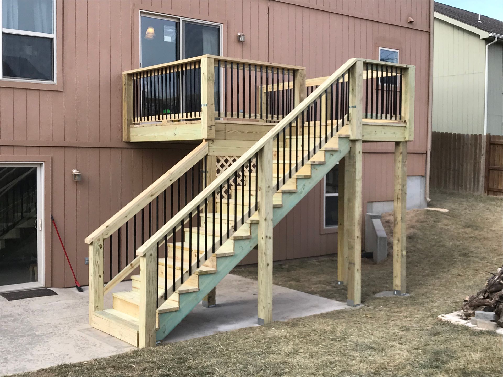 Wooden deck with stairs leading to a second-story door. Black railings and balusters. Brown siding on the house.