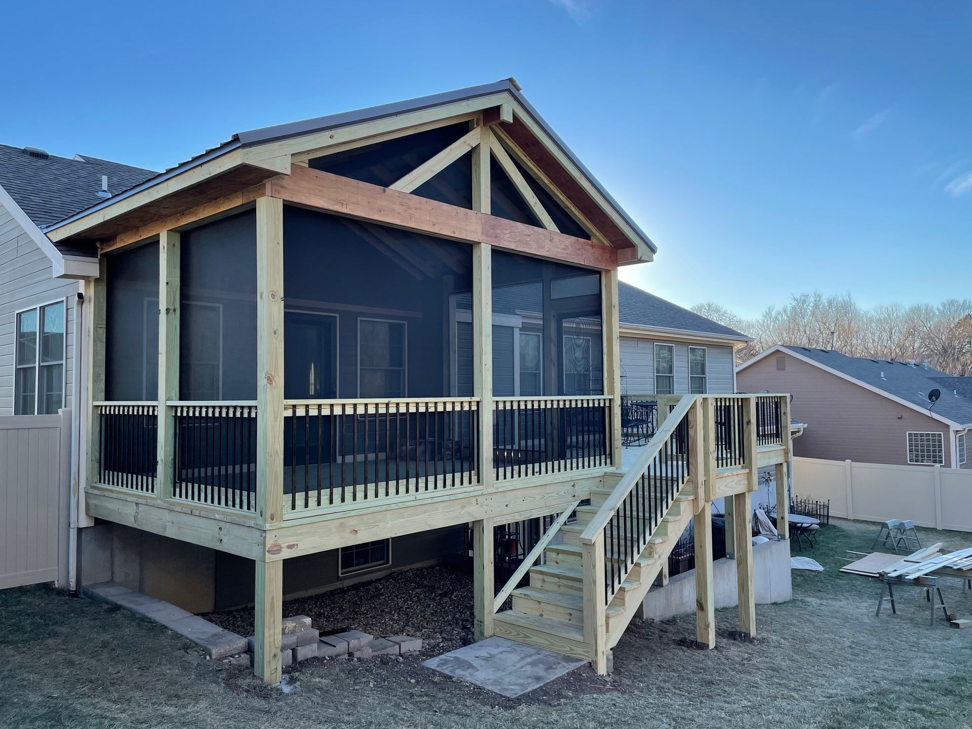 Screened-in porch and deck attached to a house with black railings and a staircase, surrounded by lawn and other houses.