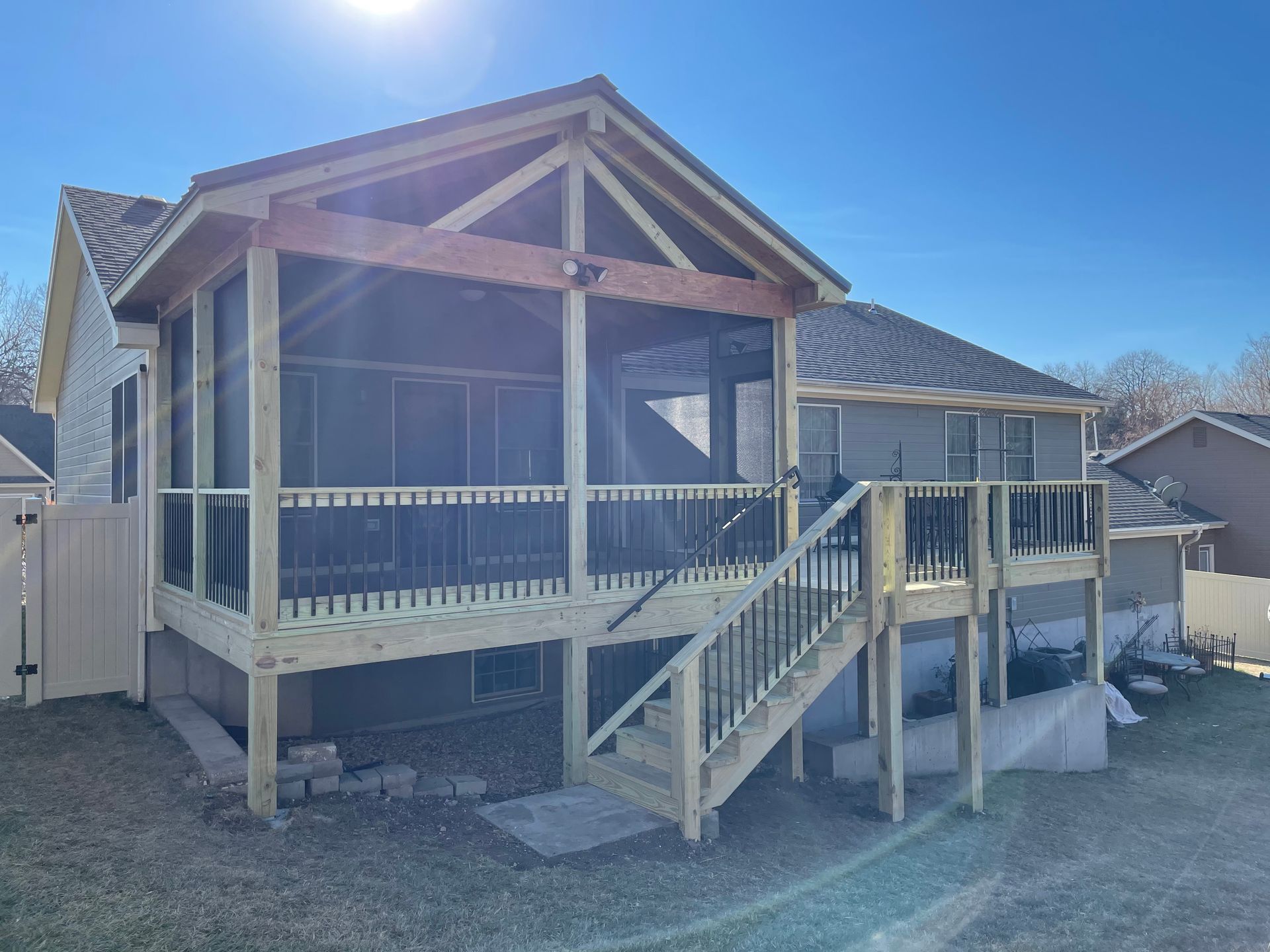 Screened porch and deck attached to a house; wooden construction; black screens and railing; blue sky.