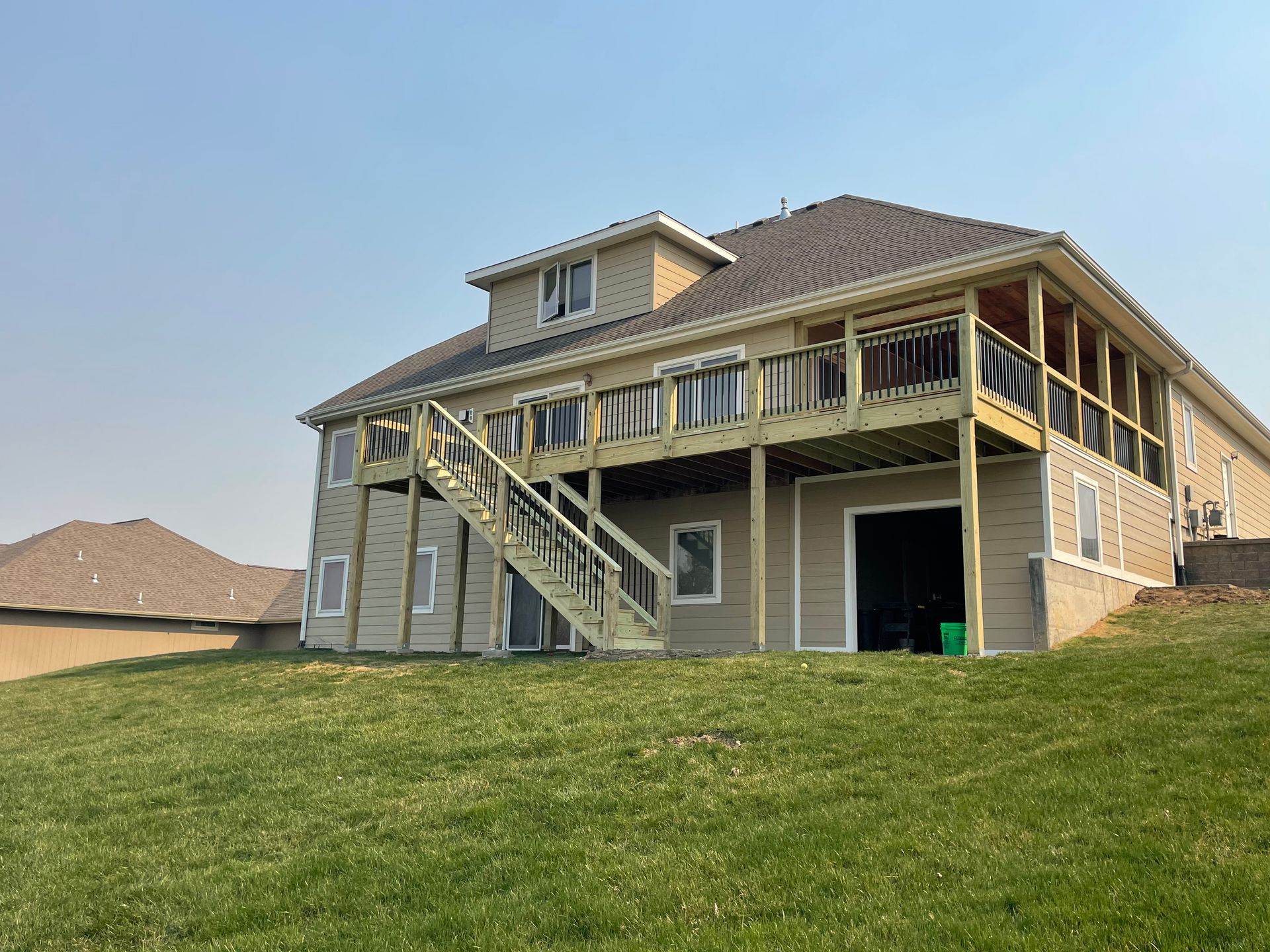 Back of beige house with wooden deck, stairs, and screened porch on a grassy hill under a blue sky.