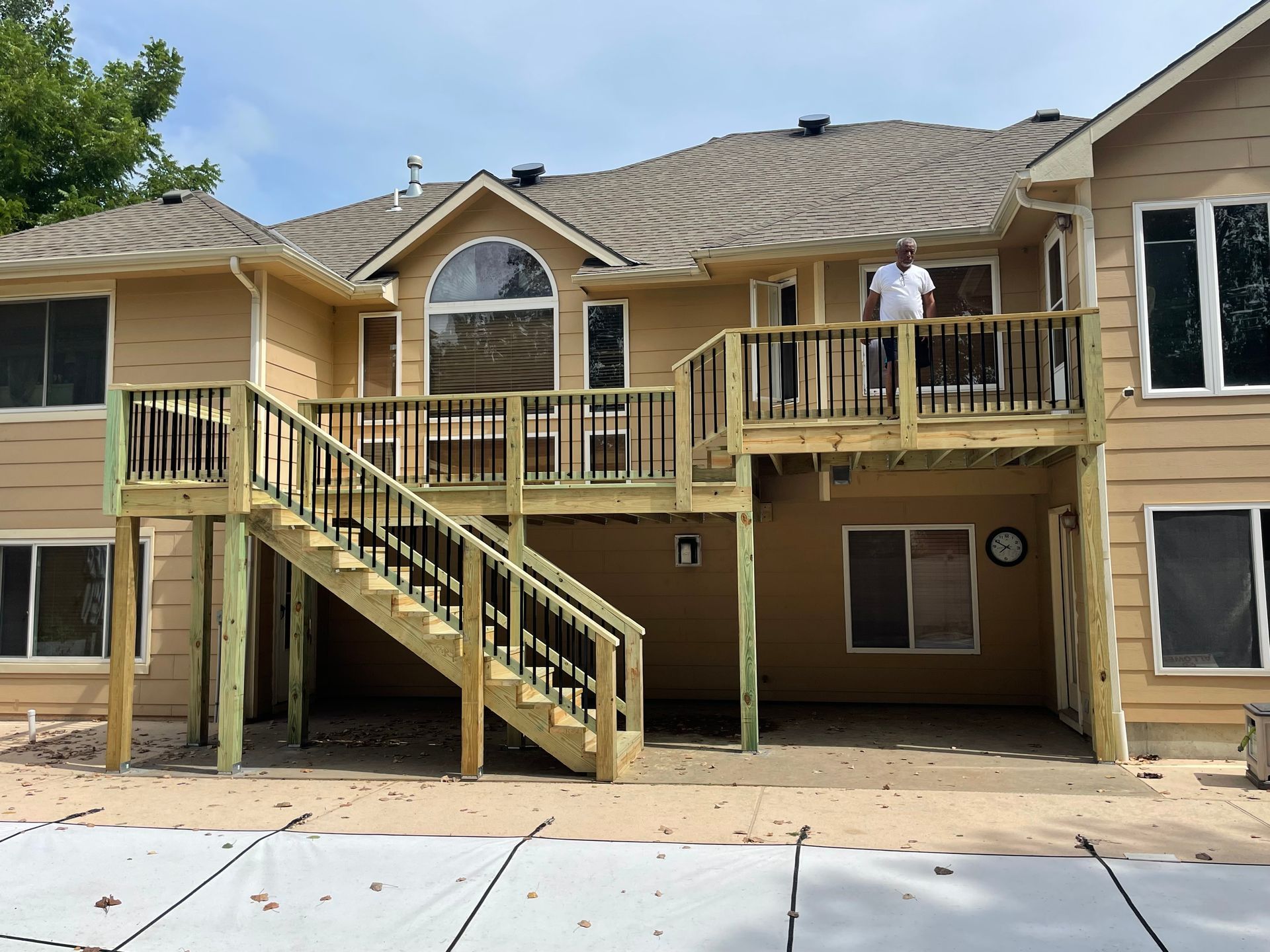 Two-level wooden deck attached to a tan house; person standing on the top level; black railings, stairs.