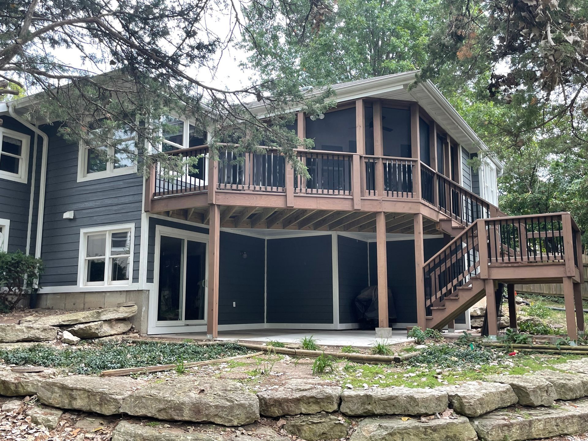 Two-story house with screened-in porch and deck, dark siding, surrounded by trees and stone retaining wall.