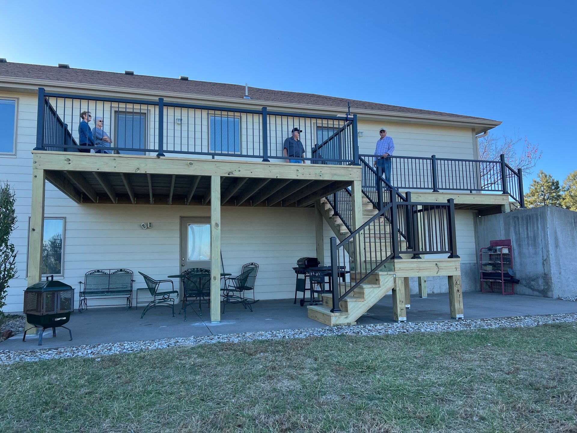 Two-story deck with people on top level; stairs descend to lower level patio with outdoor furniture.