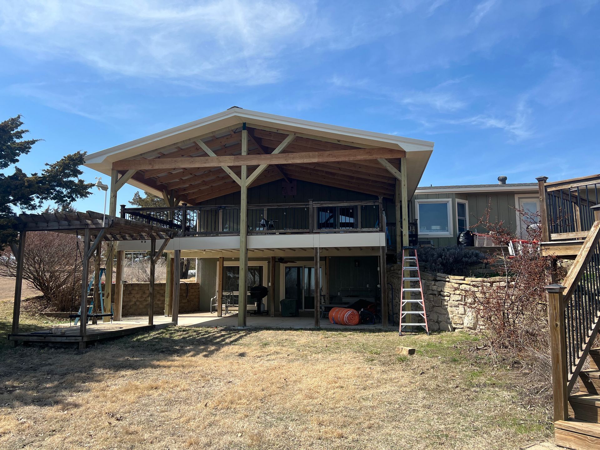 Two-story house with covered deck and pergola, overlooking water; blue sky.