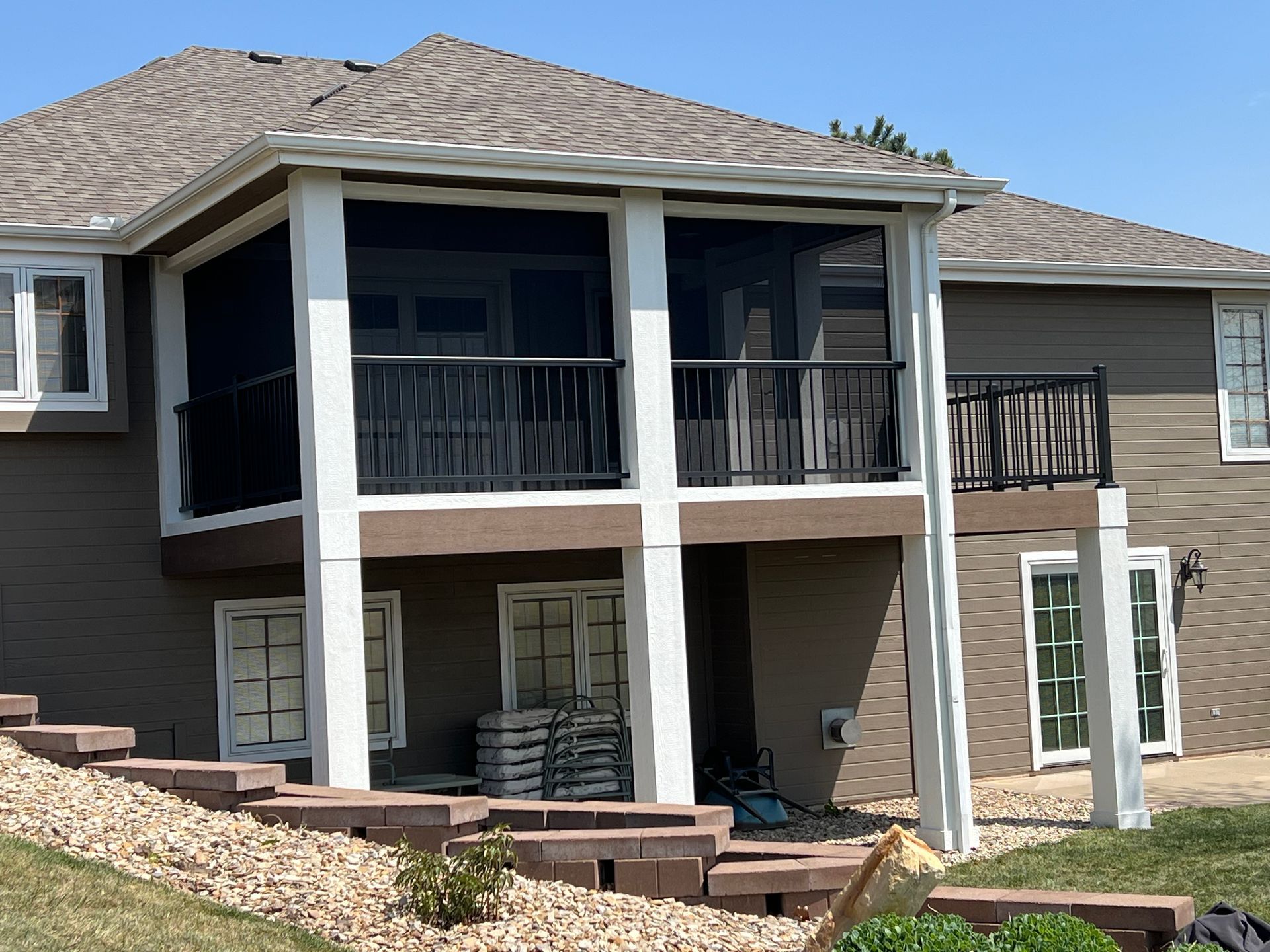 Back of a house with a screened-in porch and black railing, over a lower level with windows and support beams.