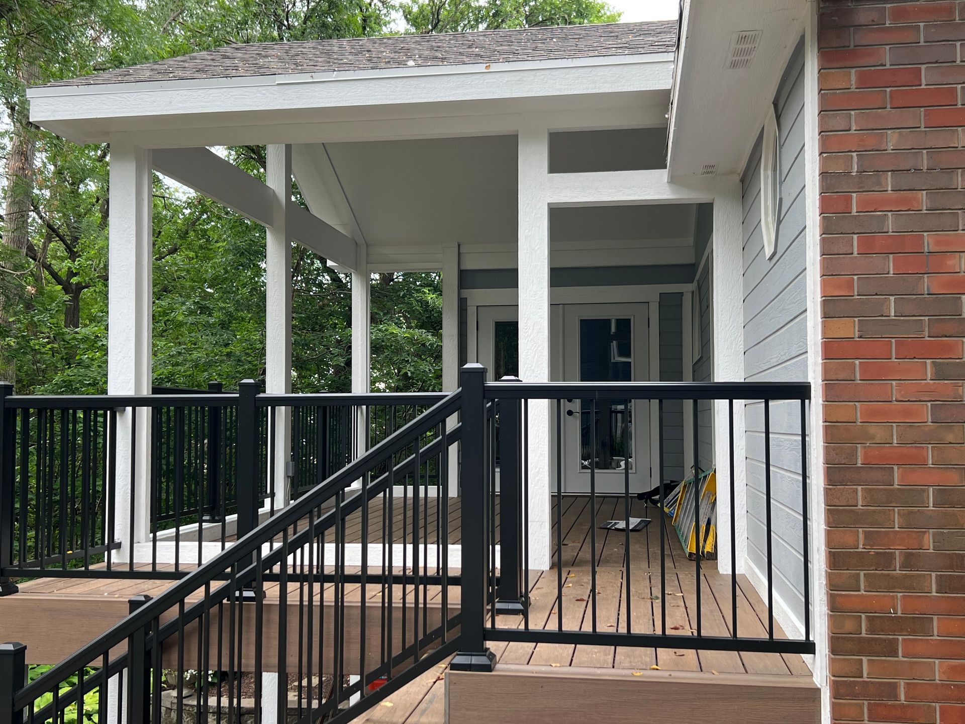 Covered porch with black railings, white columns, and gray siding. Brick wall on the right.