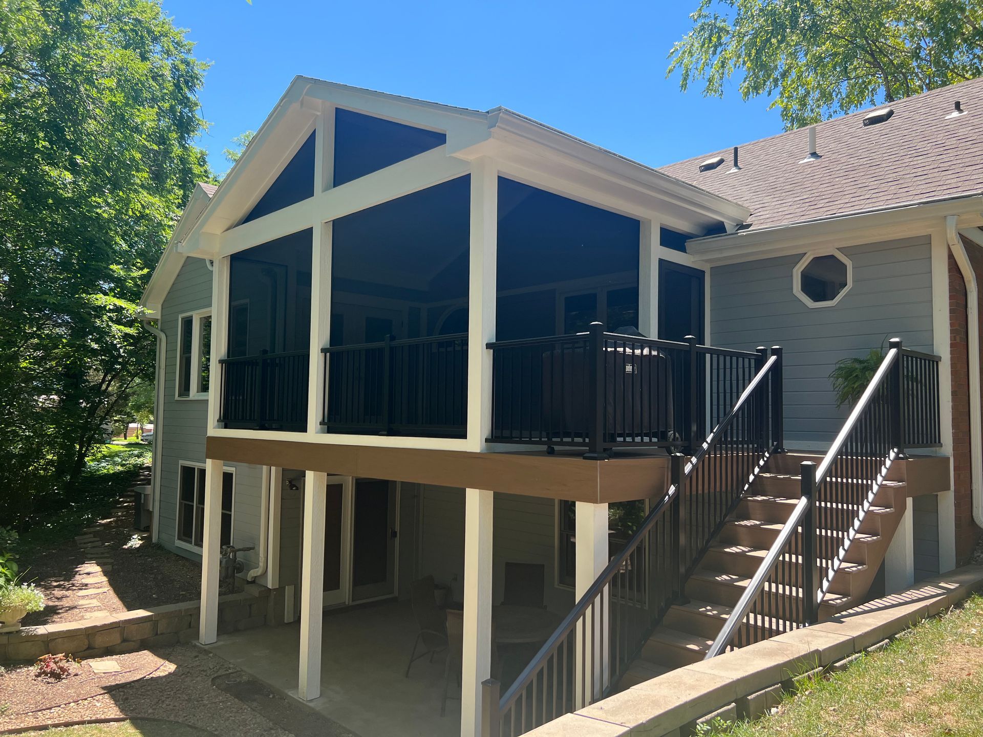 Screened-in porch addition with black railings and screens, attached to a gray house, with stairs leading down.