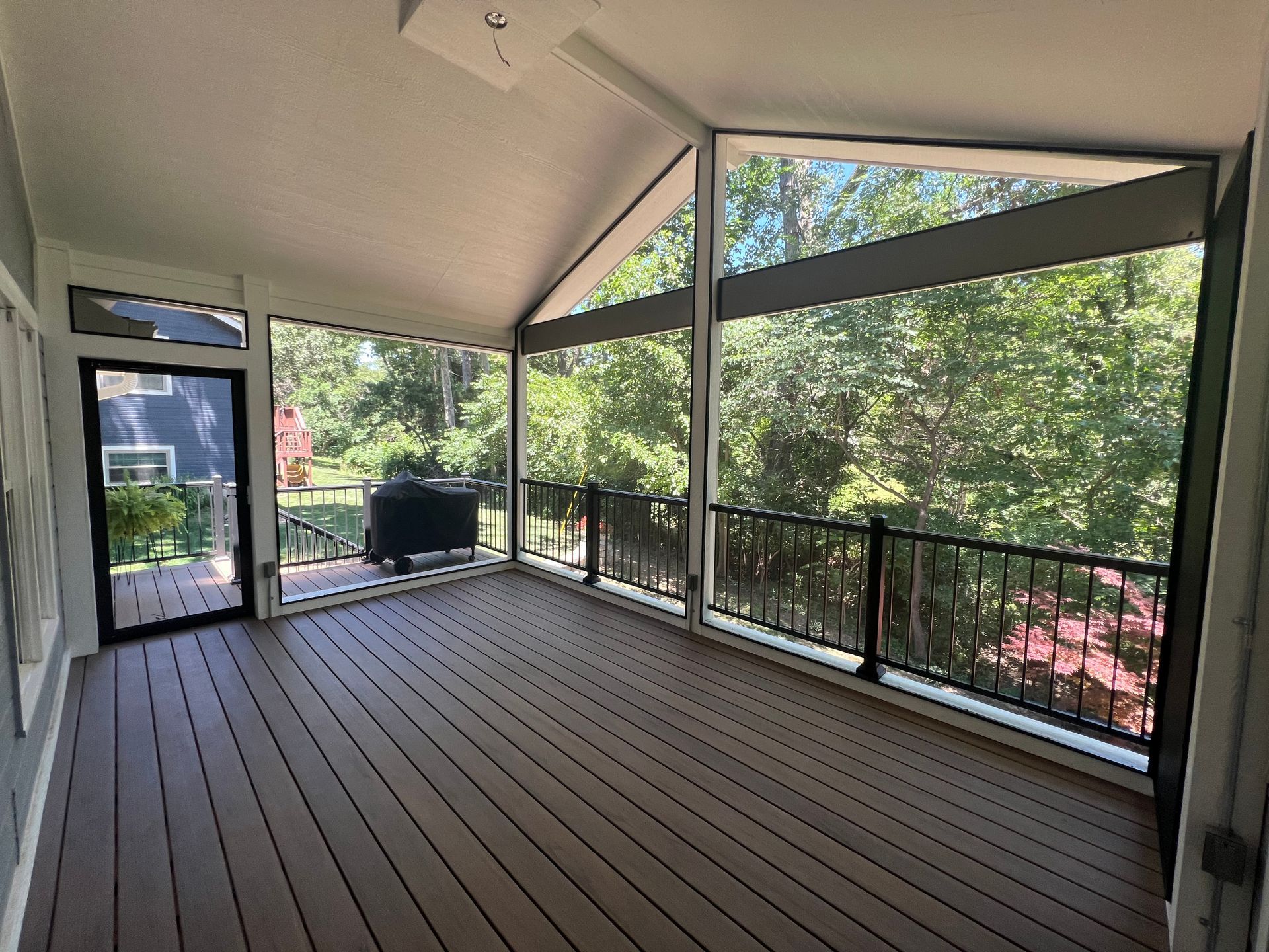Screened-in porch with brown deck, black railing, and large windows overlooking lush greenery.