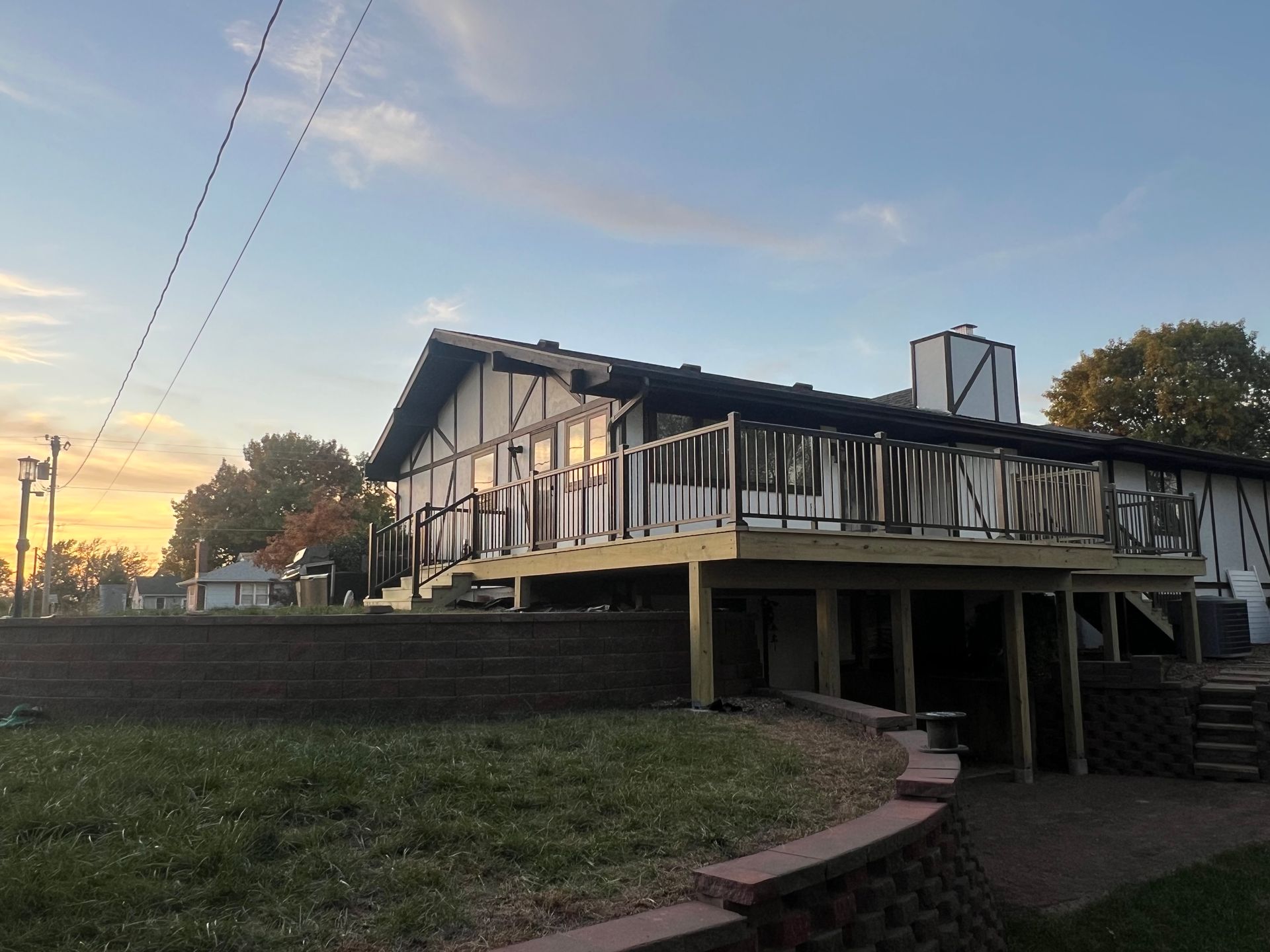 House with a black roof, deck, and white siding. A retaining wall and grass in the foreground, and a sunset in the sky.