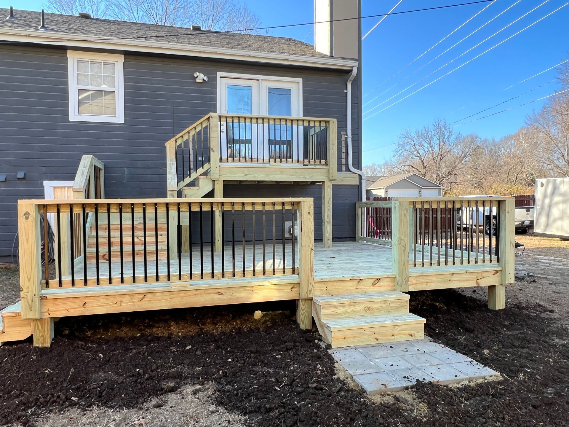 Wooden deck with stairs leading to a second level, attached to a gray house. Black railings, blue sky.