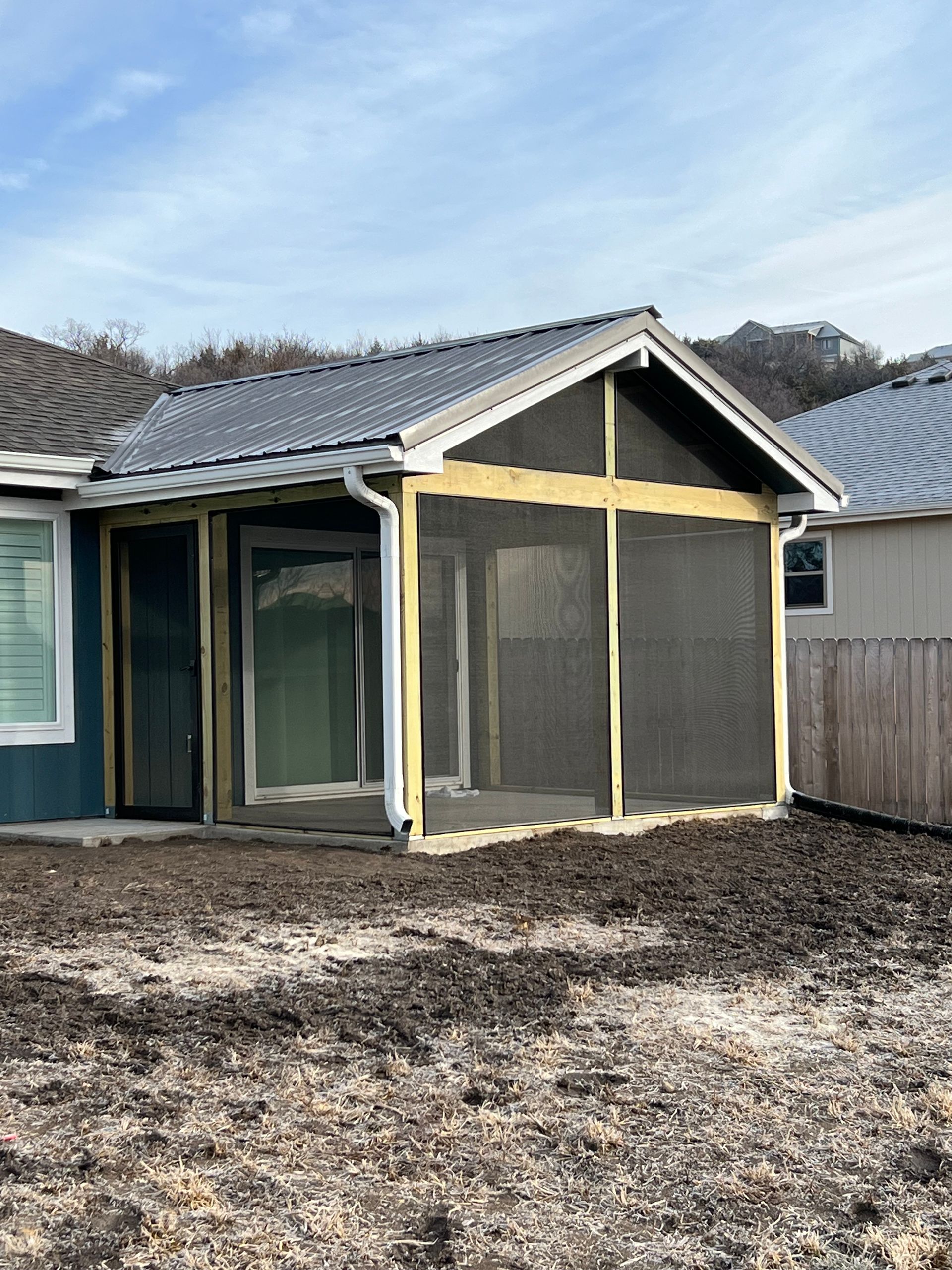 Screened porch addition on a house with a gray roof and blue siding. The porch has a concrete base and is surrounded by a yard.