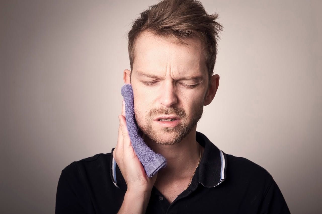Man holding a cloth to his cheek, grimacing in pain against a plain background
