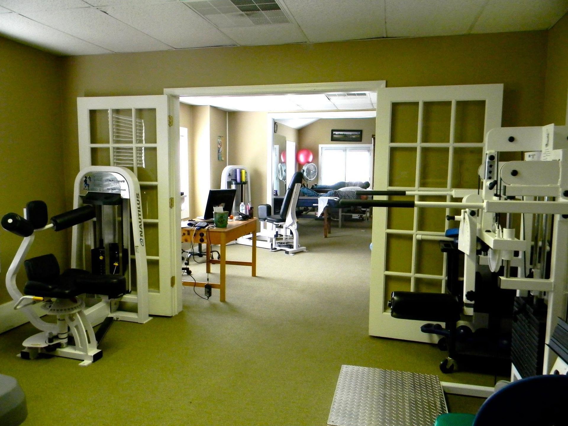 Interior of a gym with exercise machines, green carpet, and open double doors to a training room.