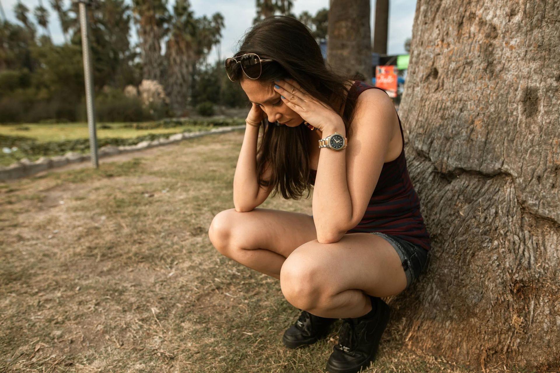 Woman crouching by a tree in a park, head in hands, wearing a black dress and boots.
