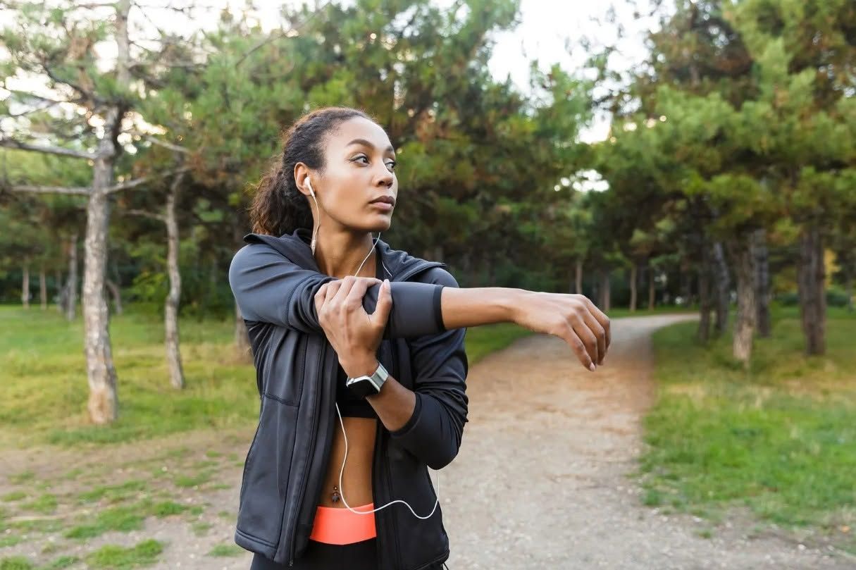 Woman stretching her arm on a park path, wearing a black jacket and coral top.