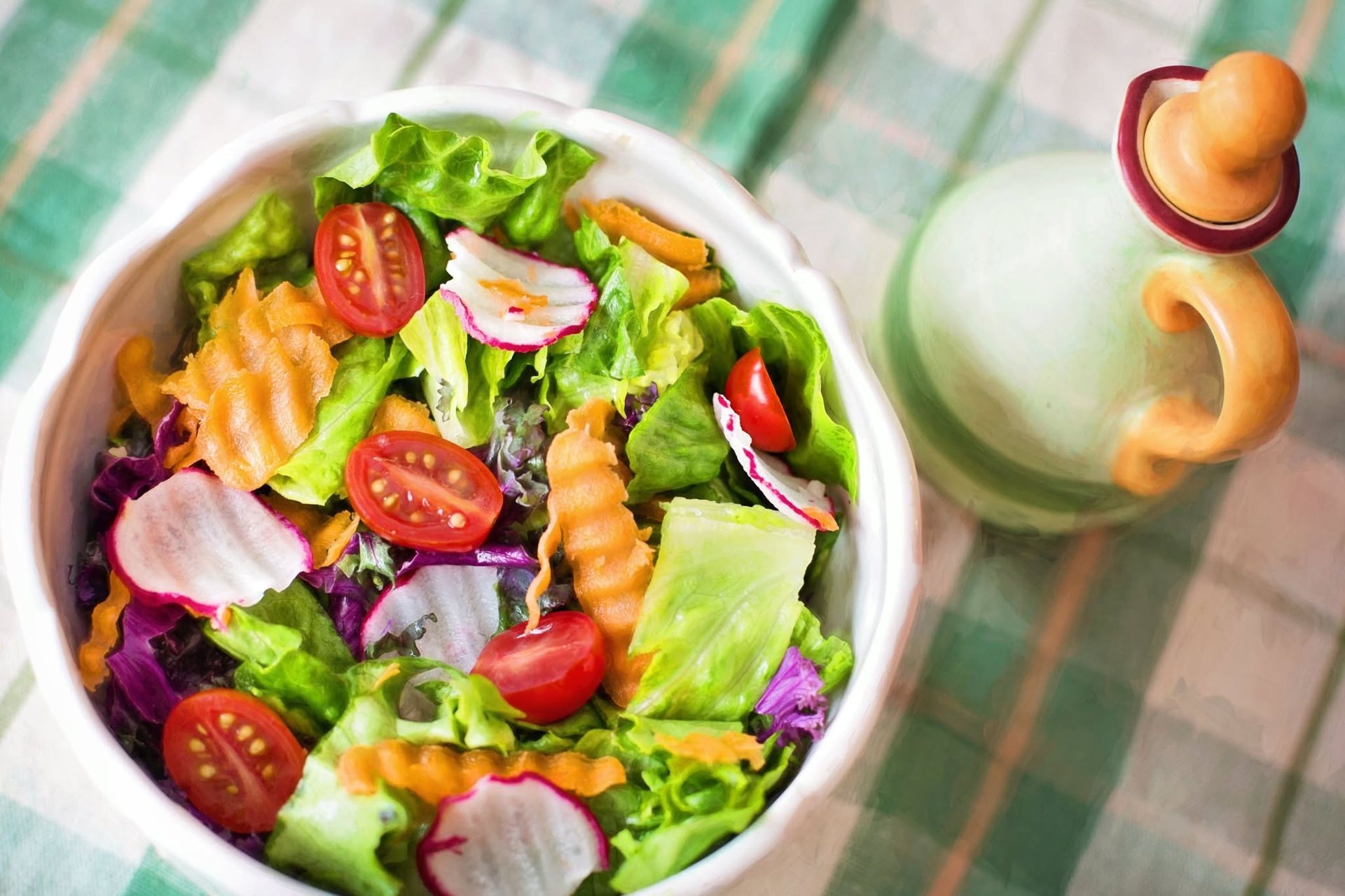 Fresh salad with cherry tomatoes, radish slices, and crinkle-cut carrots in a white bowl on a checkered cloth.