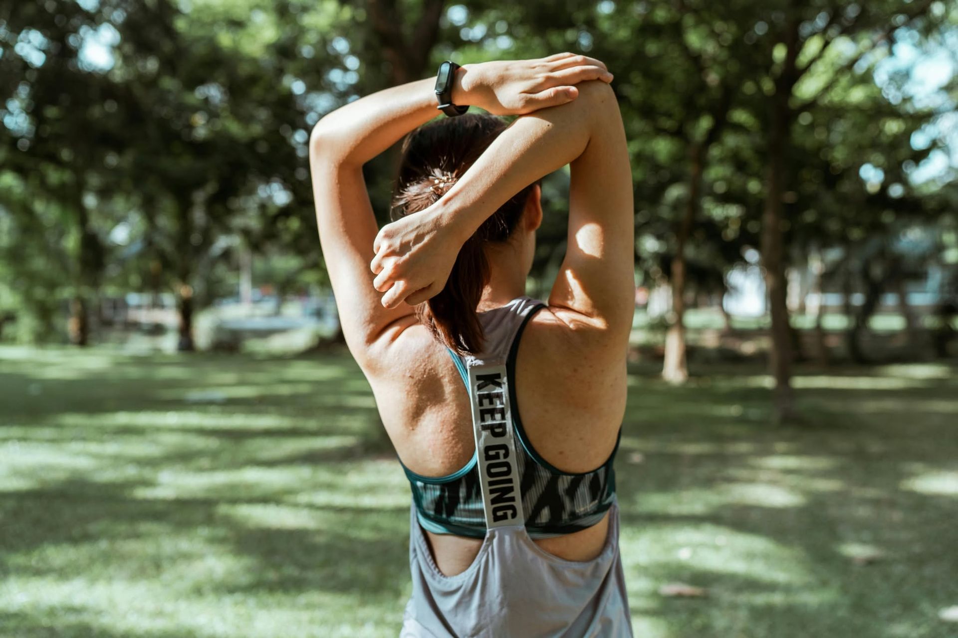 Person stretching arms overhead in a park, seen from behind, wearing a tank top.
