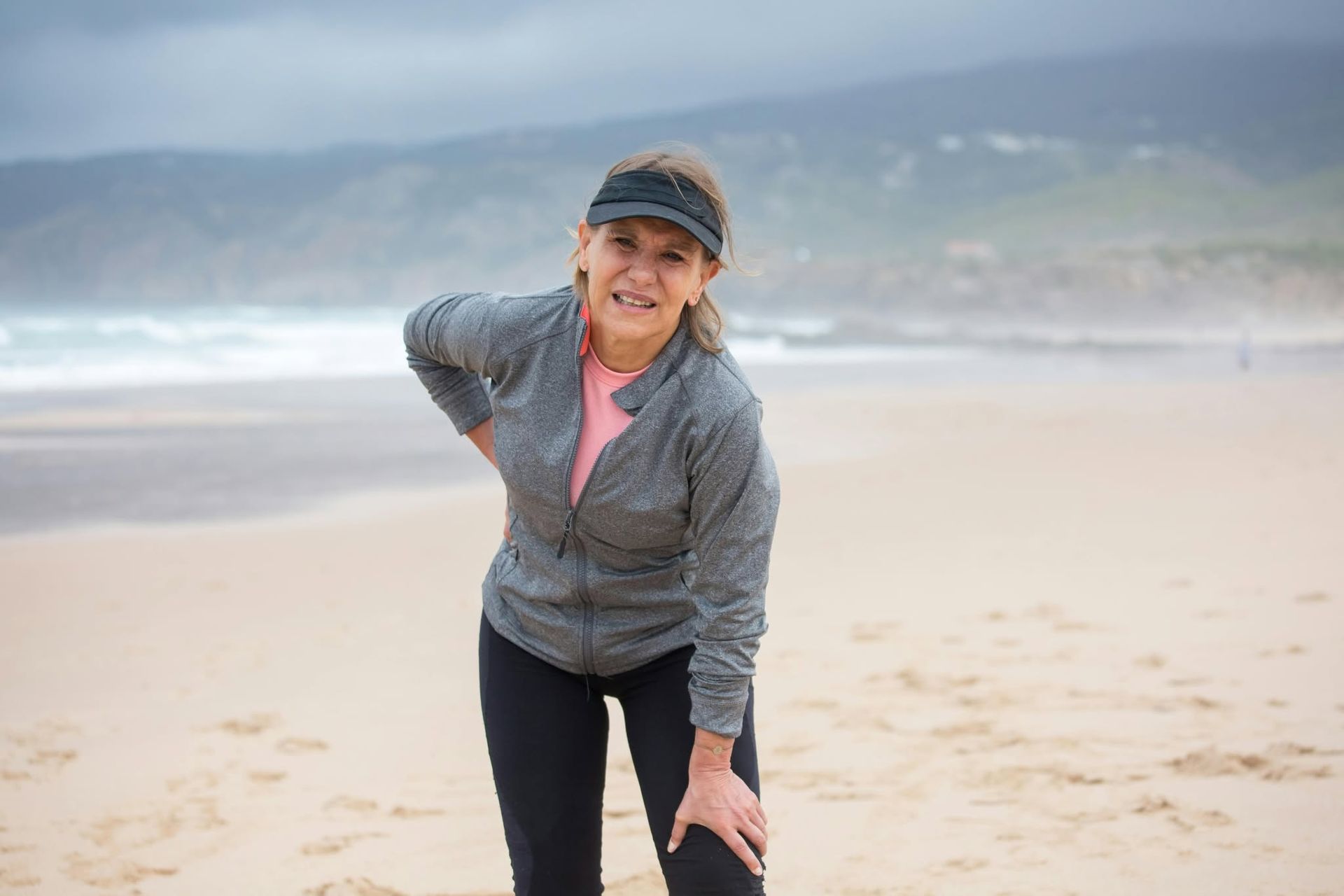 Smiling woman in a gray jacket and cap stands on a windy beach with ocean waves behind her